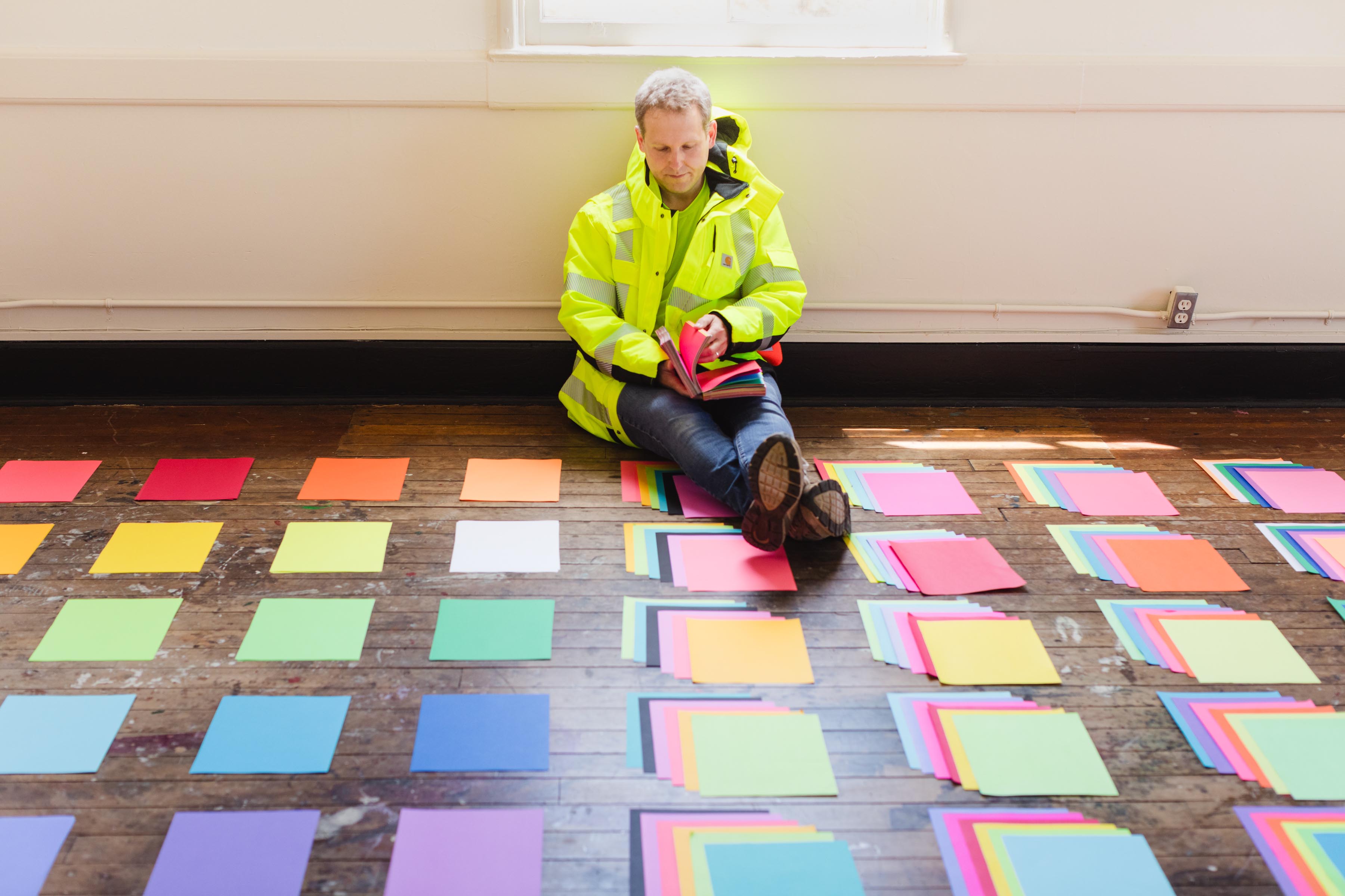 A person wearing a limegreen, high-visibility jacket sits on the floor. The person is flipping-through a book of colorful neon papers. Small stacks of colorful neon papers cover the floor.